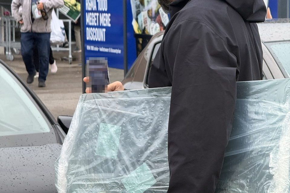 RUDEFIX-tekniker bærer en indpakket ny forrude hen til en bil på en parkeringsplads